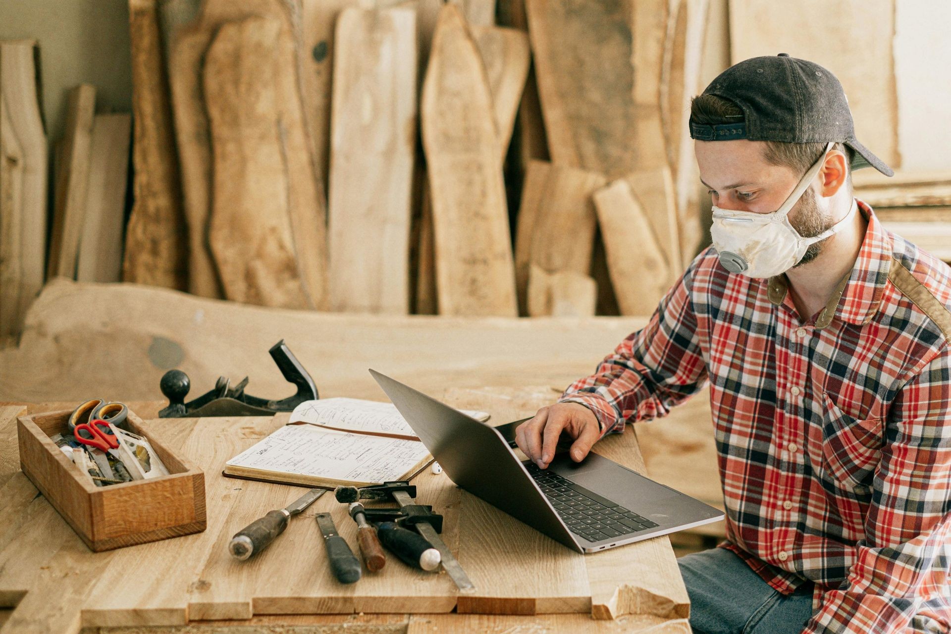 Person working on a laptop in a woodworking workshop with various tools and materials on a wooden table.