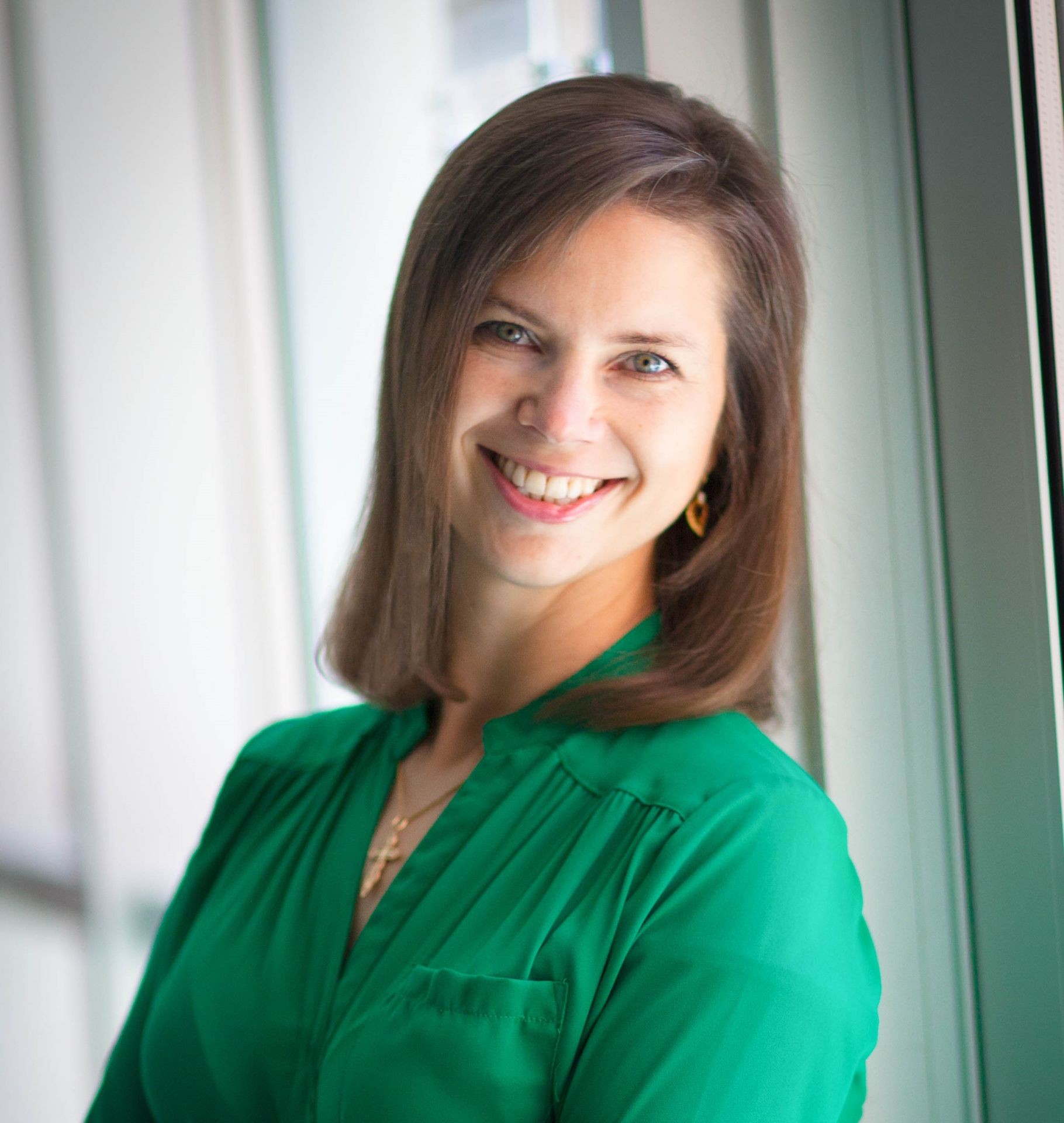 Woman with shoulder-length brown hair wearing a green top, smiling and leaning against a window.