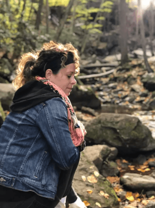 Person in a denim jacket and scarf sitting by a forest stream with rocks and autumn leaves.